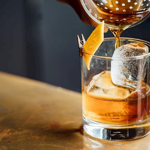 A drink being poured into a glass with ice and an orange twist, on a table, using a strainer.