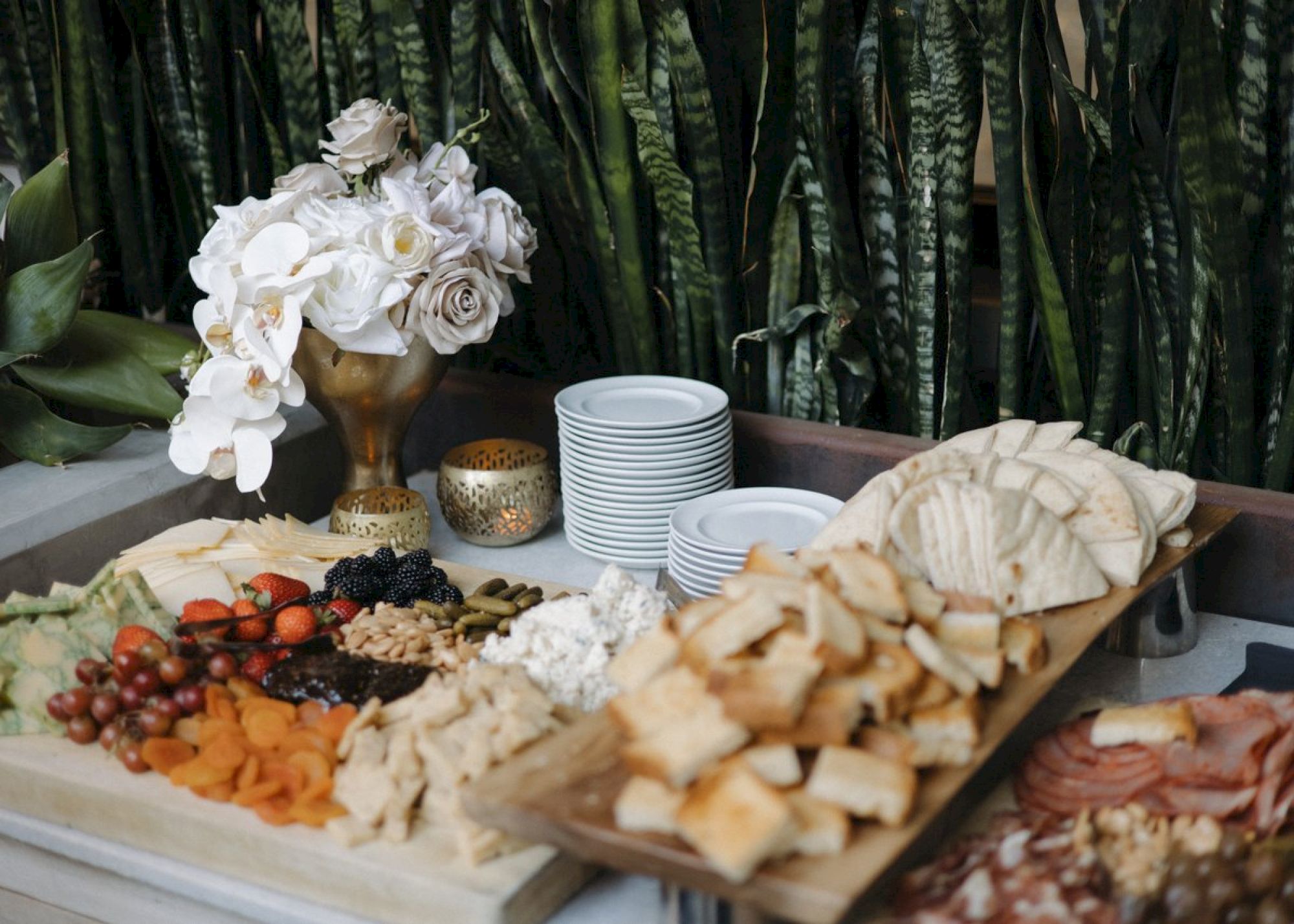 A charcuterie board with cheeses, fruits, and bread next to stacked plates, flowers, and candles.