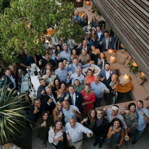A large group of people gathered outdoors, posing for a photo. They are surrounded by greenery and patio furniture.