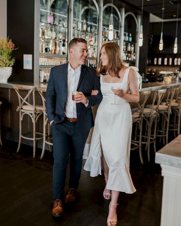A couple dressed elegantly, holding drinks, smiles at each other while walking in a stylish bar or restaurant setting.