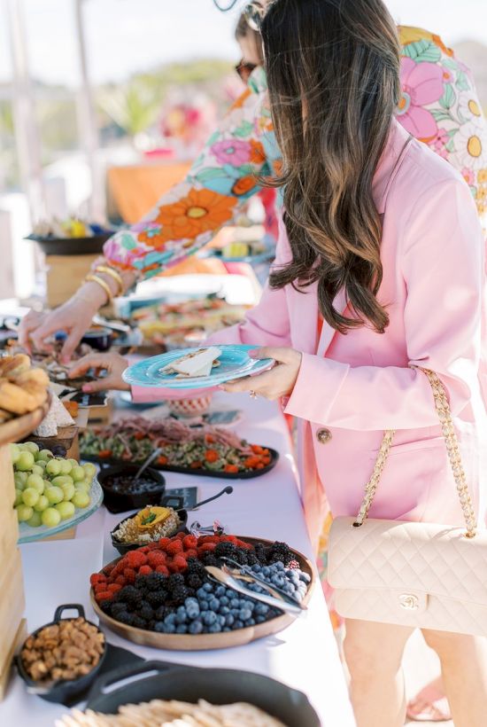 A woman in a pink outfit is selecting food from a buffet table with various fruits and dishes, while others also pick food items.