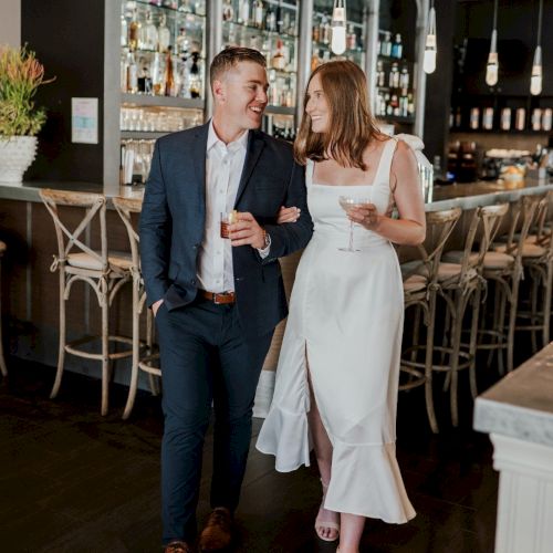 A couple is walking arm-in-arm in a stylish bar. The man wears a suit; the woman is in a white dress. They both hold drinks, smiling.