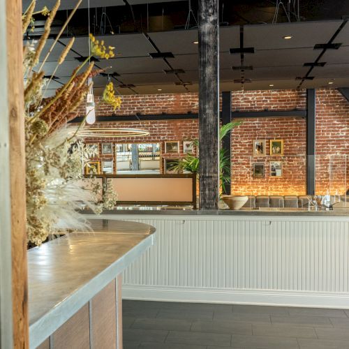 The image shows a stylish interior with a counter, dried plants, exposed bricks, and modern lighting, creating a cozy atmosphere.