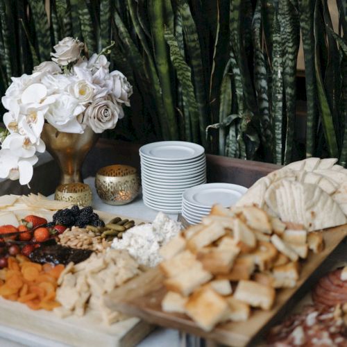 A table with a variety of cheeses, fruits, bread, and pastries. Decor includes white flowers and plates stacked nearby.