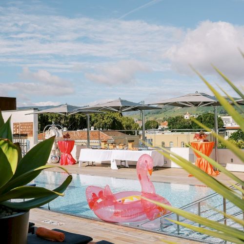 A rooftop pool scene with inflatable flamingos, plants, and tables set up, against a backdrop of mountains and a partly cloudy sky.