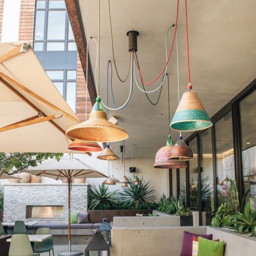 An outdoor seating area with wicker pendant lights, vibrant cushions, umbrellas, and potted plants, under a modern architectural space.