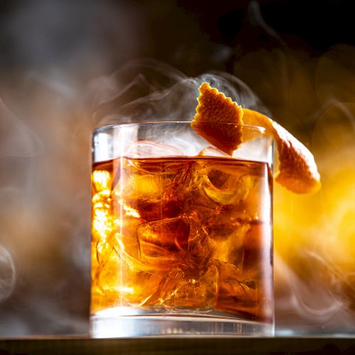 A glass of an amber-colored cocktail with ice and a citrus garnish, surrounded by a smoky atmosphere, creating a dramatic presentation.