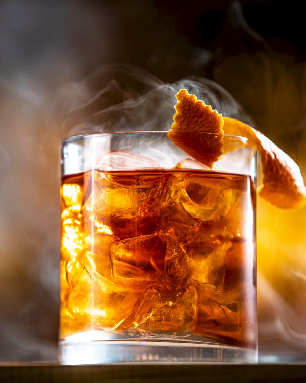 A glass of an amber-colored cocktail with ice and a citrus garnish, surrounded by a smoky atmosphere, creating a dramatic presentation.