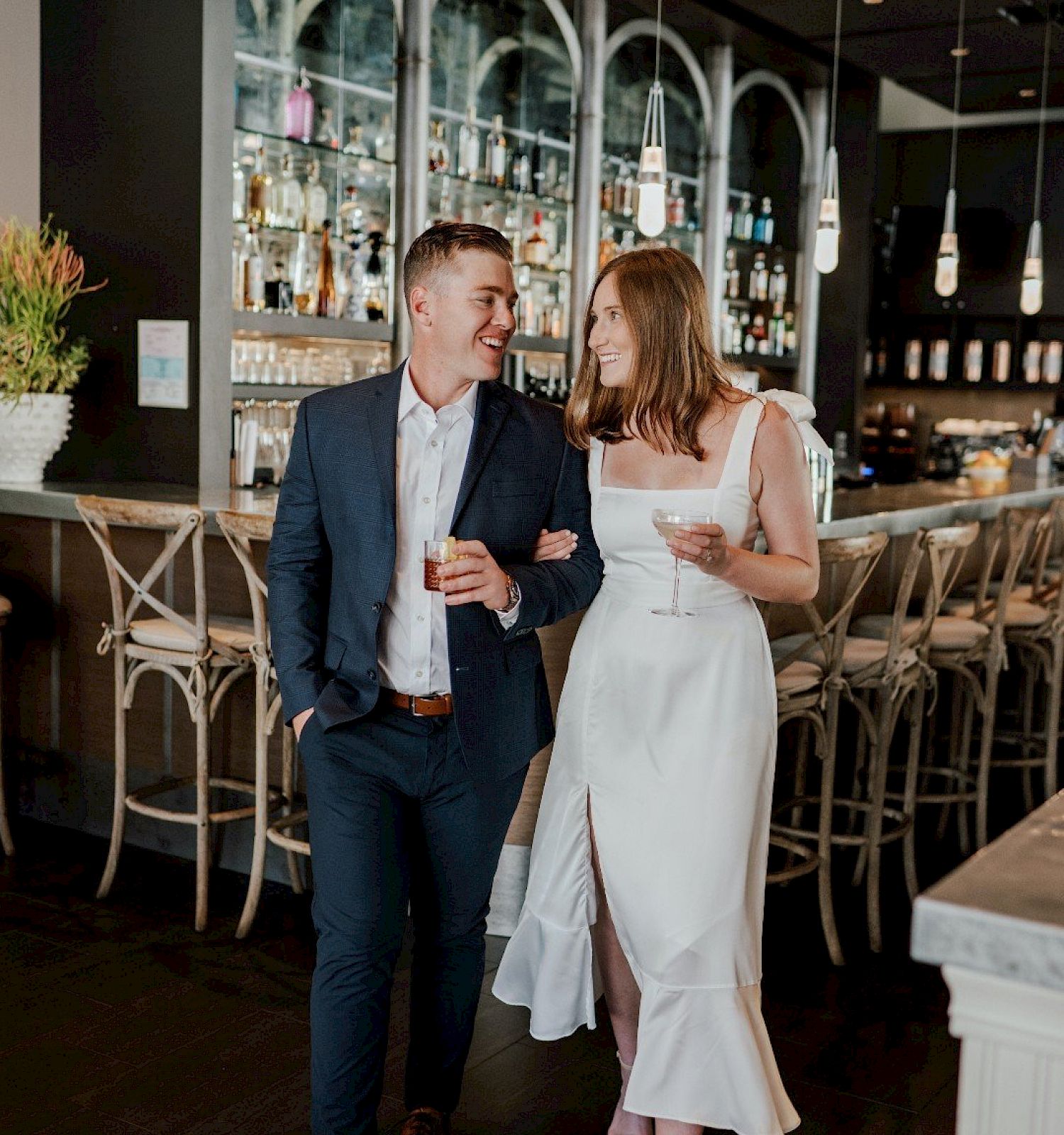 A couple is walking through a stylish bar. The man is in a suit, and the woman wears a white dress, both holding drinks.