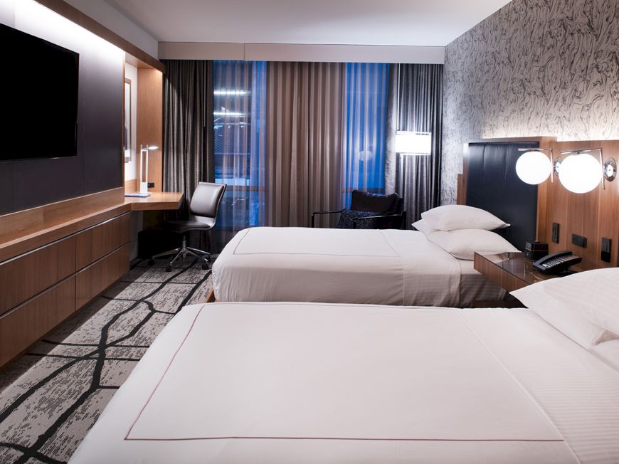 The image shows a modern hotel room with two beds, a flat-screen TV, a desk, and lamps, featuring neutral tones and contemporary decor.