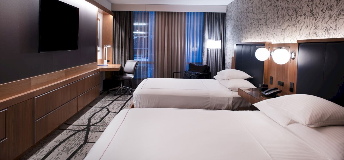 The image shows a modern hotel room with two beds, a flat-screen TV, a desk, and lamps, featuring neutral tones and contemporary decor.