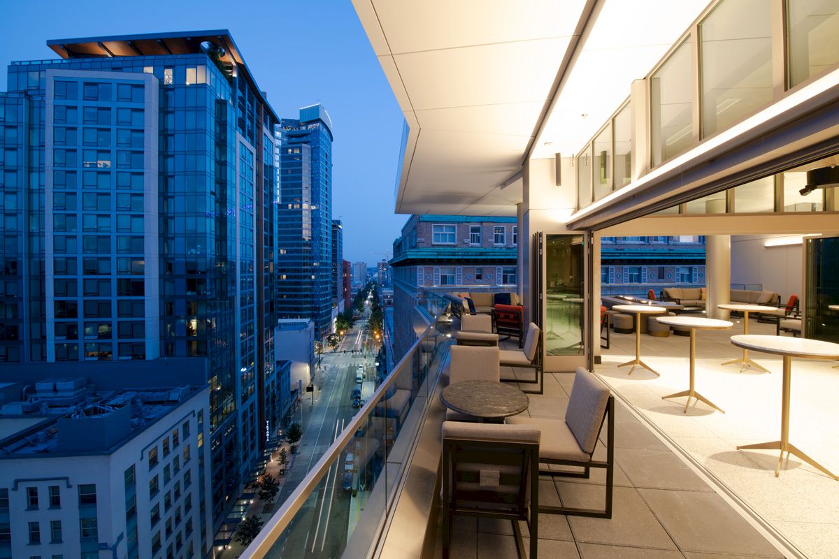 A modern rooftop terrace with chairs and tables overlooks a cityscape at dusk, featuring tall buildings and a vibrant skyline.