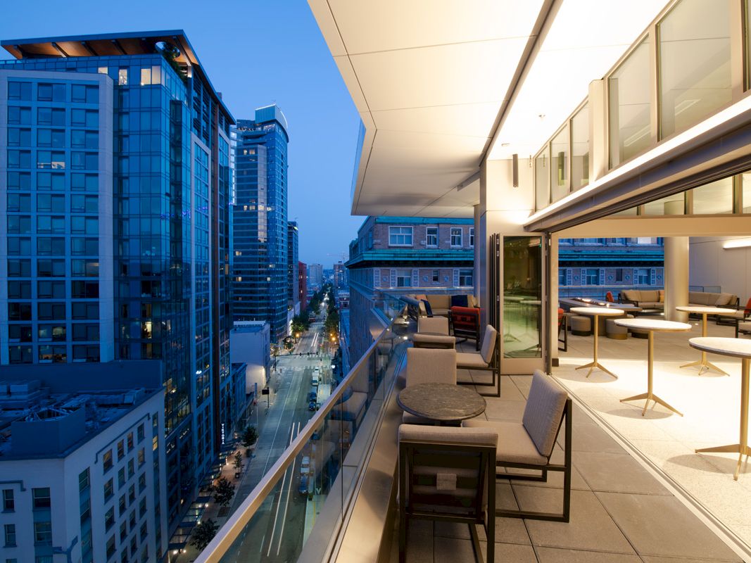 A modern rooftop terrace with chairs and tables overlooks a cityscape at dusk, featuring tall buildings and a vibrant skyline.