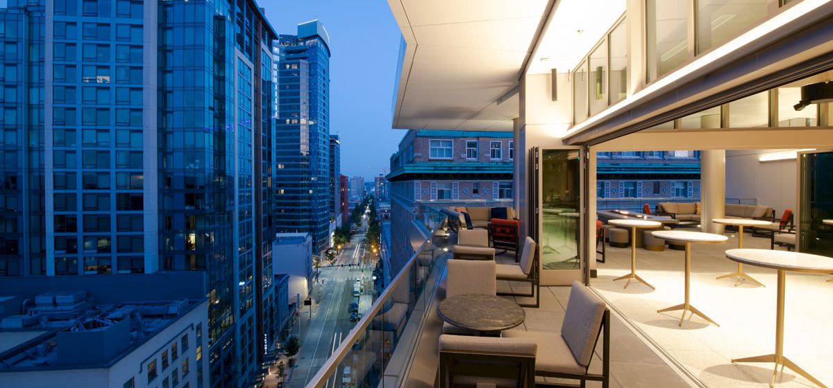 A modern rooftop terrace with chairs and tables overlooks a cityscape at dusk, featuring tall buildings and a vibrant skyline.