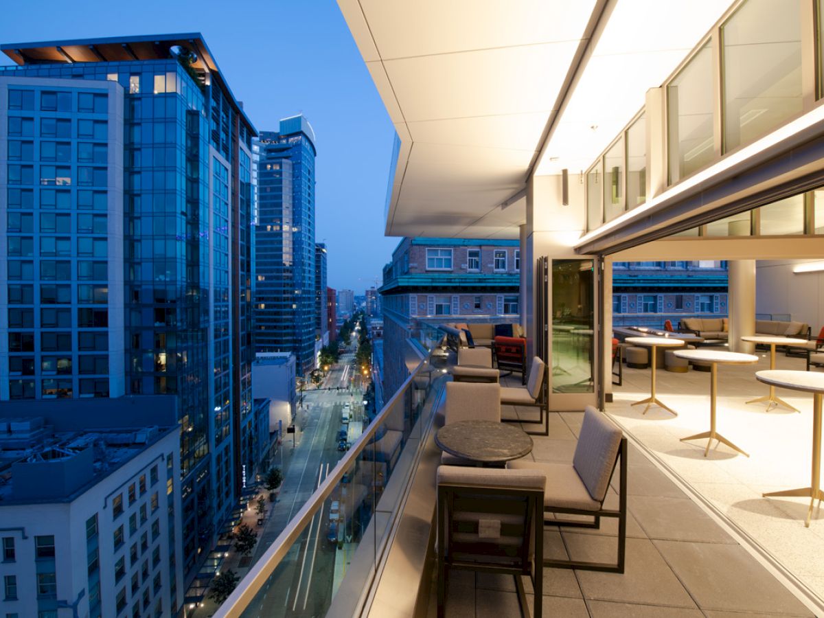 A modern rooftop terrace overlooks a cityscape at dusk, with cozy seating areas and lit buildings in view.