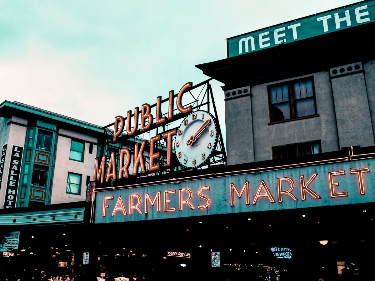 The image shows a neon sign for a public farmers market, featuring a clock and surrounded by buildings and a "Meet the…" phrase.