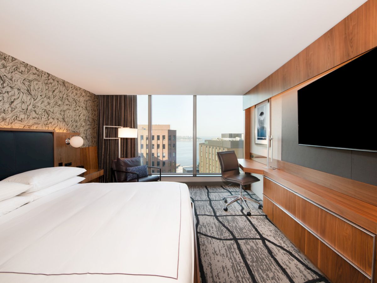 A modern hotel room featuring a large bed, flat-screen TV, desk, and a window with a city view, decorated in neutral tones.