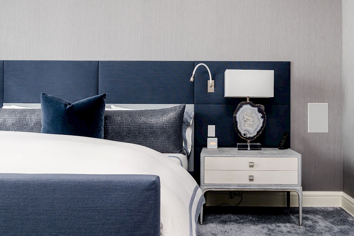 A modern bedroom with a blue and white color scheme, featuring a bed, side table, lamp, and decorative accents on a gray carpet.