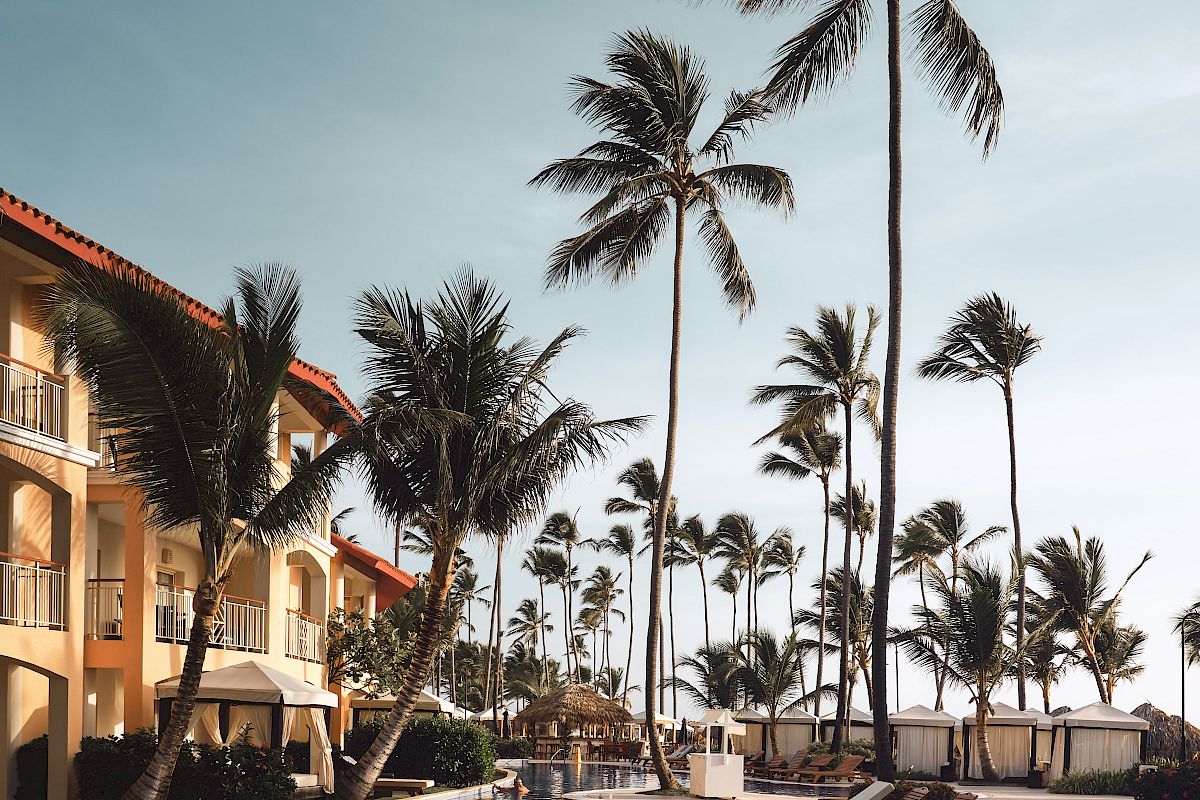 The image shows a tropical resort scene with a pool, lounge chairs, a building, and palm trees under a clear sky.