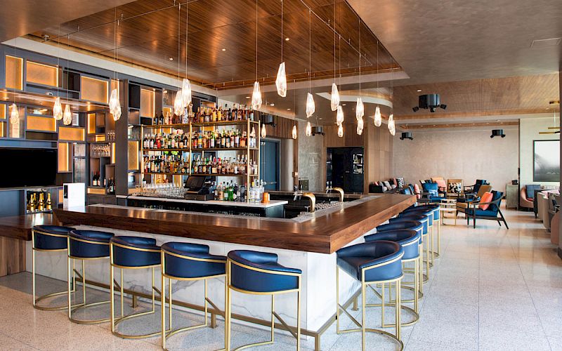The image shows a modern bar with blue stools, a stocked bar, pendant lights, and lounge area seating.