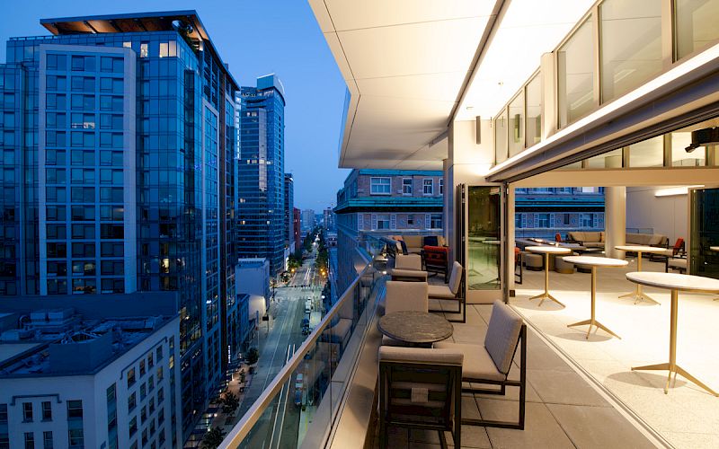 A rooftop terrace view shows modern city buildings during twilight, with seating areas and tables set along the edge of the terrace.